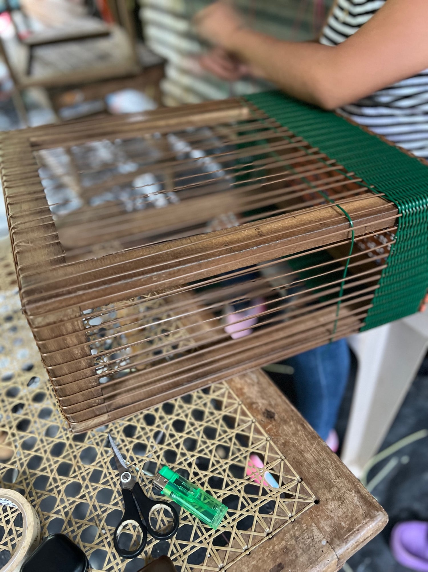 Person weaving green thread on a wooden frame, with scissors and a green lighter on the table beside.