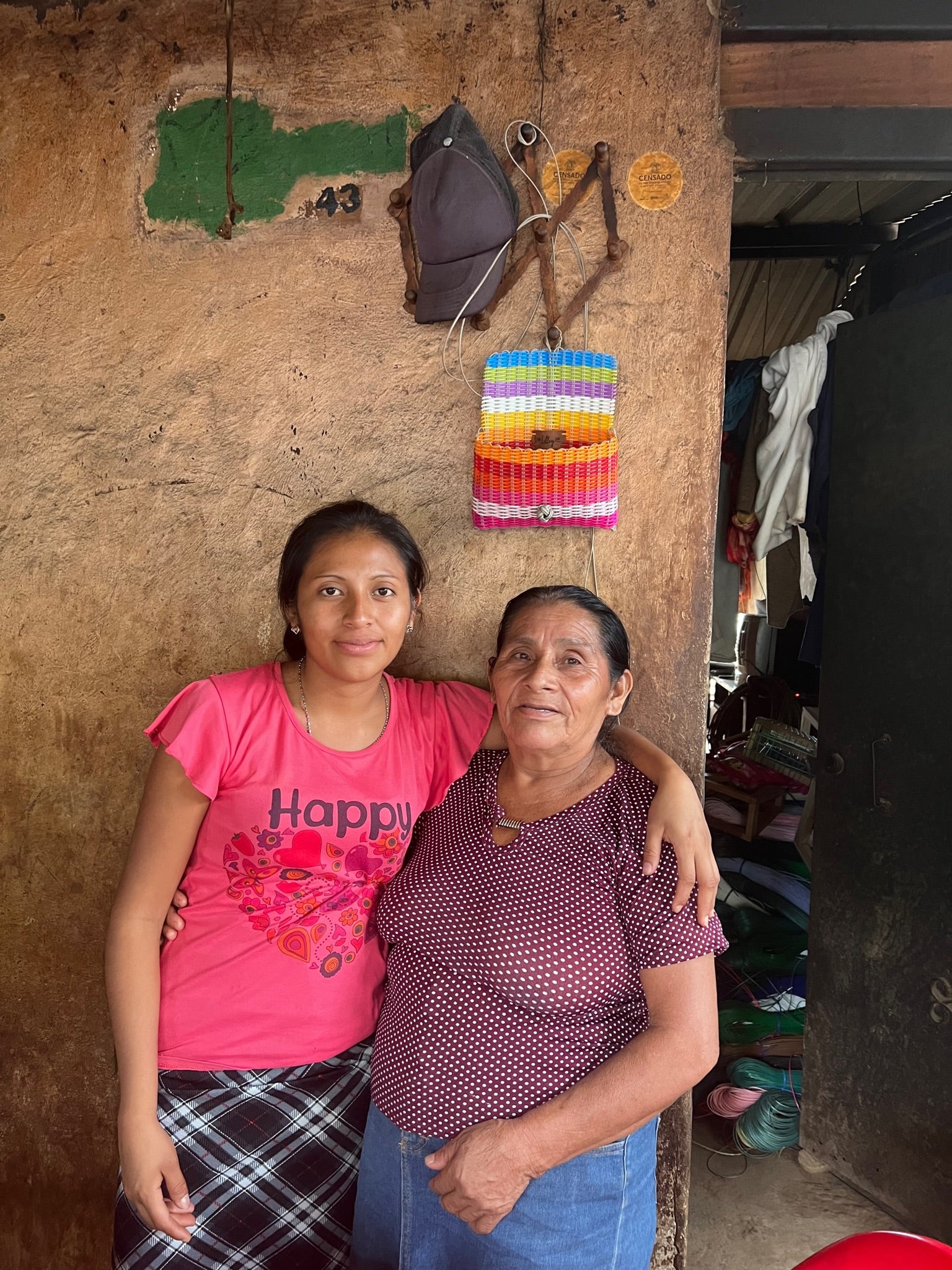Two women standing in front of a colorful wall adorned with various objects, smiling and dressed in casual clothing.
