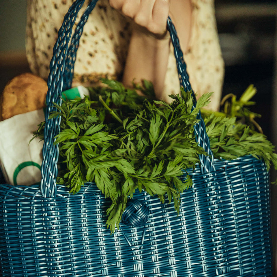 A woman holds a blue woven basket overflowing with fresh veggies and assorted items, showcasing her harvest.