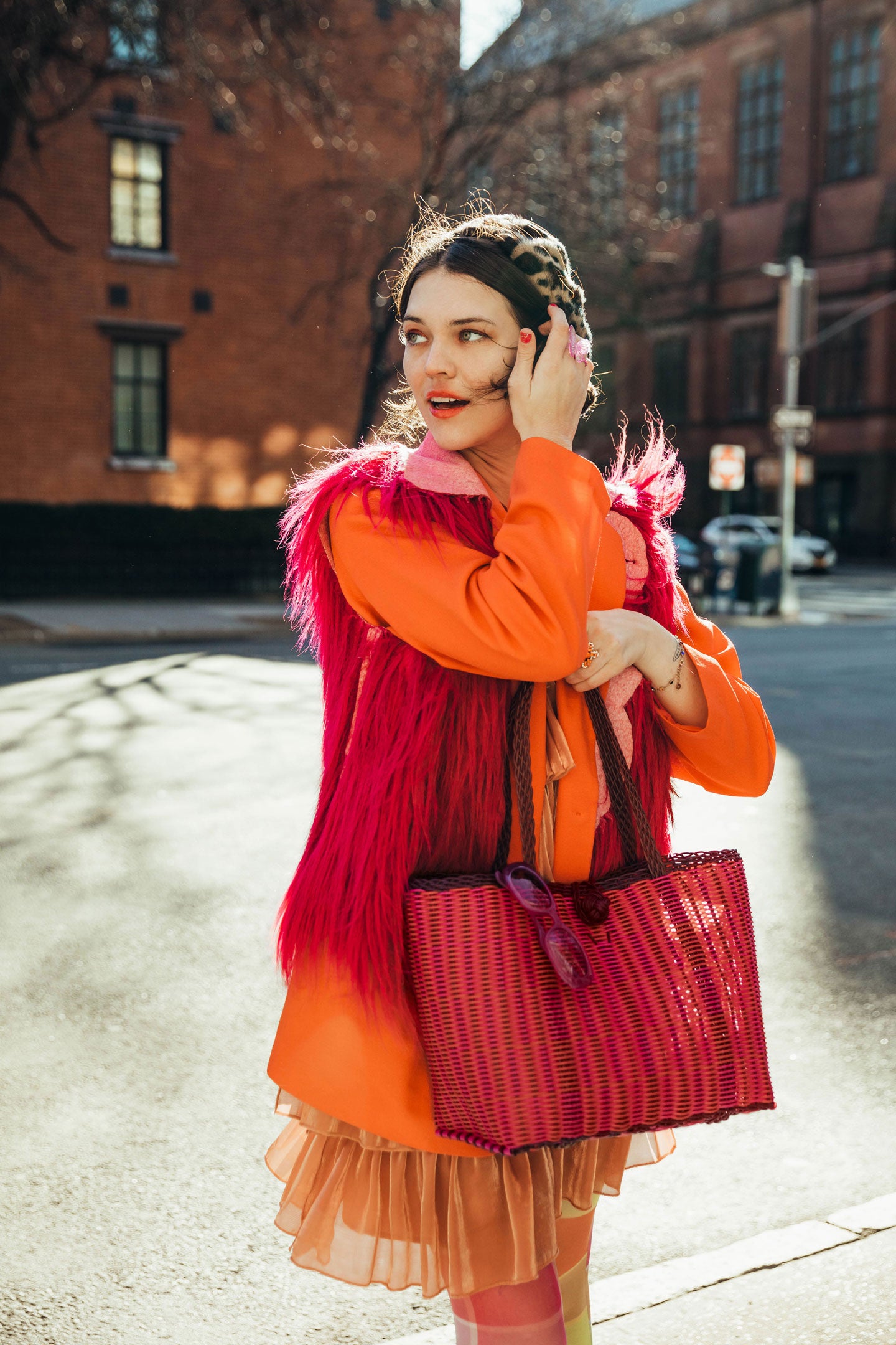 A woman in a colorful outfit stands on a city street, holding a woven bag. She wears an orange coat with a pink fur vest and looks to the side. Sunlight casts shadows on the ground.