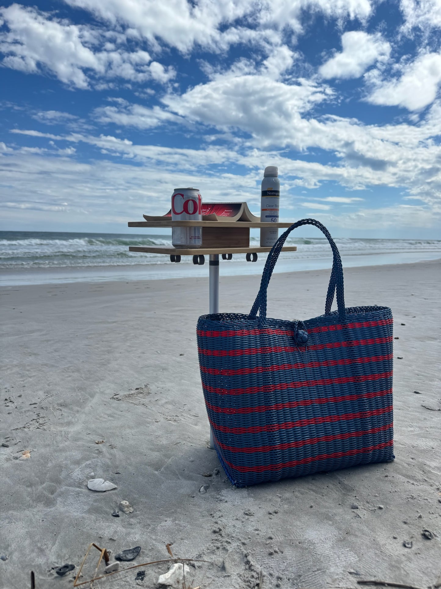 Blue and red striped bag on a sandy beach with a blue sky and clouds.