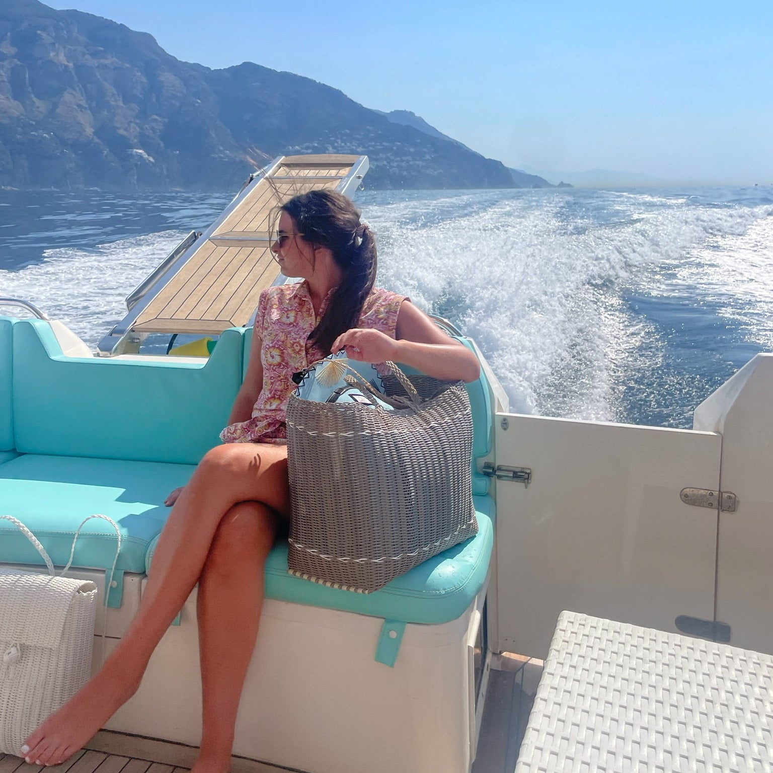 A woman sits on a turquoise and cream motorboat deck, holding a large gray hand-woven Lilley Line tote bag. The boat is cruising across the Amalfi coastline in the background under a clear blue sky.
