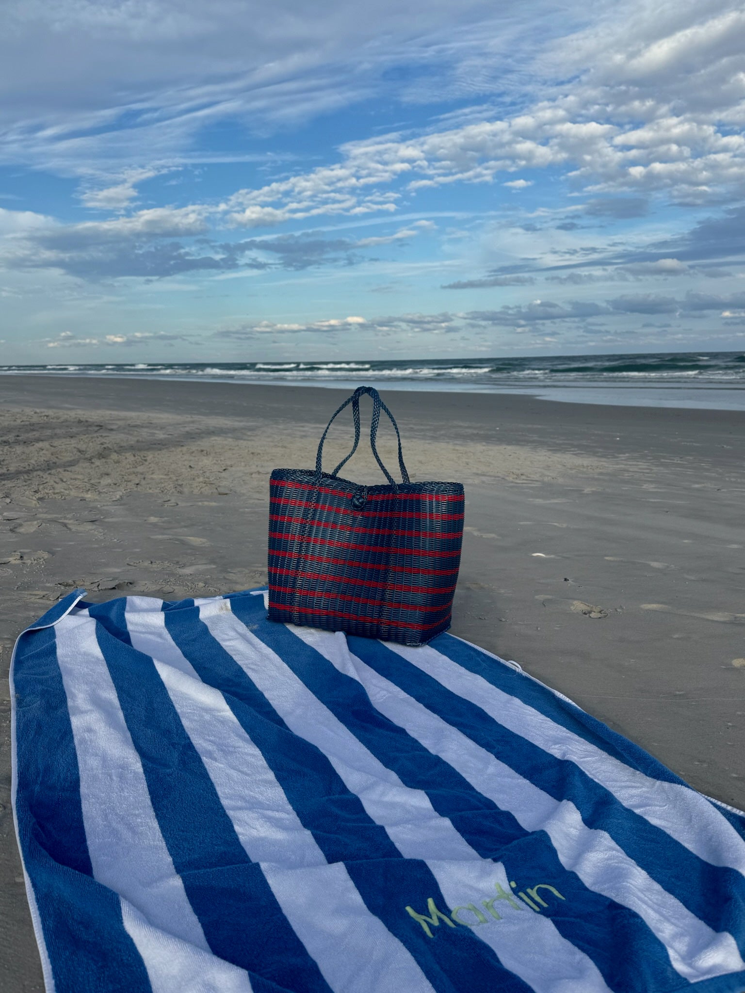 Blue and red striped bag on a blue and white striped towel at the beach with ocean and sky in the background.