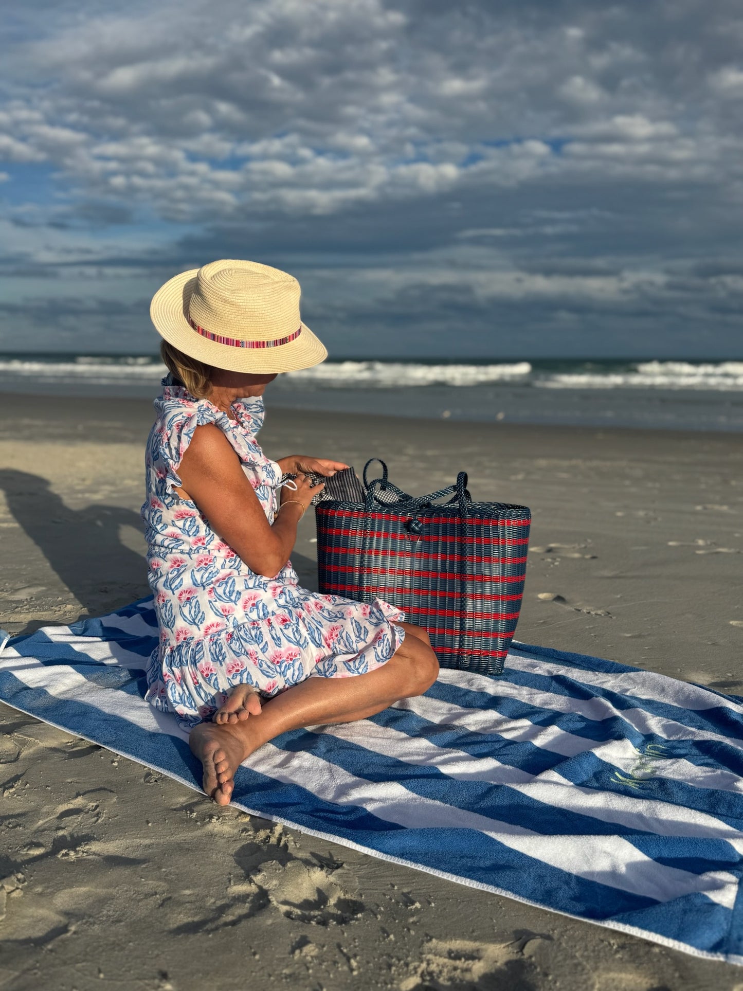 Person sitting on a beach with a blue and white striped towel and a red and blue bag.
